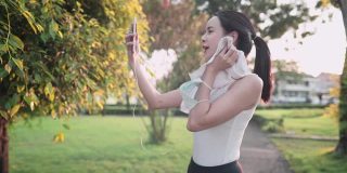 年轻漂亮的女人在运动服装擦拭脸上的汗水，通过智能手机视频通话在公园锻炼