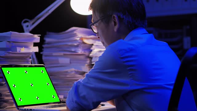 57岁的亚洲高级男子看着绿色屏幕在远程地点通过电脑在家里工作到深夜。绿屏新技术概念。