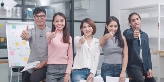 一群亚洲年轻的创意人士在时髦的休闲穿着微笑和大拇指在创意办公室的工作场所。不同的亚洲男性和女性站在一起创业。