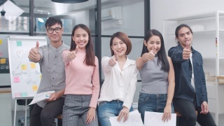 一群亚洲年轻的创意人士在时髦的休闲穿着微笑和大拇指在创意办公室的工作场所。不同的亚洲男性和女性站在一起创业。视频素材模板下载