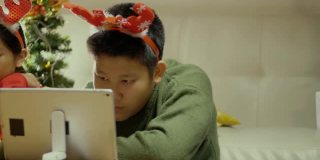 快乐的亚洲儿童穿着毛衣和发带使用智能手机和平板电脑在晚上一起在家里以圣诞装饰为背景，假日概念。