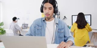 新常态下的亚洲商人社交距离为病毒预防在家庭办公室工作时使用笔记本电脑向同事介绍视频通话计划。