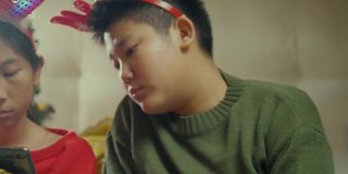快乐的亚洲儿童穿着毛衣和发带使用智能手机和平板电脑在晚上一起在家里以圣诞装饰为背景，假日概念。