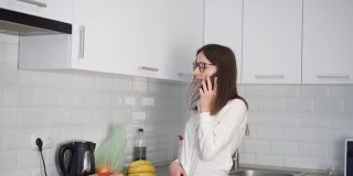 一位戴着眼镜、心情愉快的年轻女子在厨房边吃着食物边讲电话。