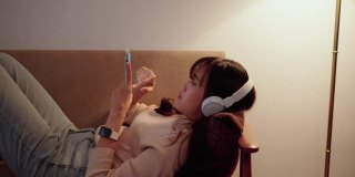 美丽的年轻亚洲女人拿着她的智能手机，正在沙发上播放音乐。