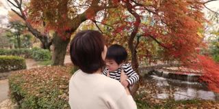 年轻的母亲在秋天的红叶下捡起她的宝贝儿子