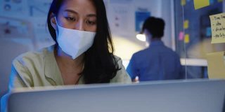 疲惫不堪的年轻亚洲女性戴着口罩用笔记本电脑工作，患有办公室综合症，颈部疼痛，在办公室加班。