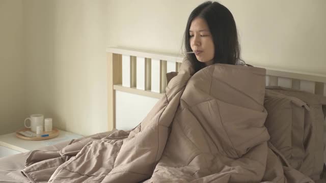 生病的亚洲妇女坐在床上，盖着毯子，有寒冷，颤抖，用温度计测量体温，看着摄像机。