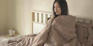 生病的亚洲妇女坐在床上，盖着毯子，有寒冷，颤抖，用温度计测量体温，看着摄像机。