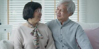 肖像快乐的资深亚洲夫妇放松坐在家里微笑在沙发沙发上看相机享受退休或隔离时间。有吸引力的亚洲成年人老年人幸福的生活方式。
