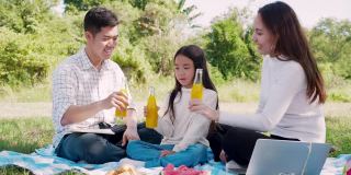 幸福的家庭野餐。亚洲父母(父亲，母亲)和小女孩喝橙汁，并有乐趣和享受自己一起在野餐布在绿色花园