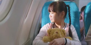 亚洲小女孩拥抱熊娃娃的肖像，坐在飞机上。孩子感到兴奋和快乐的旅行与她的家人。乘客在飞机上的运输概念。