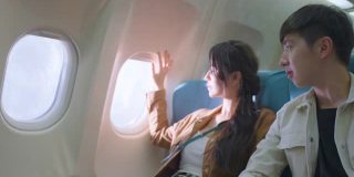 一对亚洲年轻夫妇坐在靠窗的座位上，看着窗外，指着窗外。男人和女人感到兴奋和幸福的蜜月甜蜜旅行旅行的飞机运输。