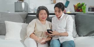亚洲家庭妇女和她的母亲使用智能手机教玩互联网一起在客厅在家里的幸福和微笑。两代沟技术使用生活方式的概念。