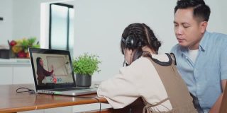 由于冠状病毒大流行，亚洲小女孩通过数字远程互联网会议从学校老师那里学习在线课程。孩子看着电脑笔记本电脑屏幕坐在父亲。