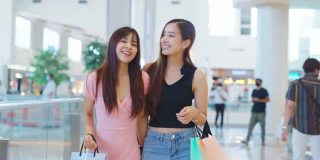 两个快乐的亚洲年轻女性在商场室内购物在销售季节在年底。女孩们拿着购物袋，快乐地走着，微笑着一起买东西