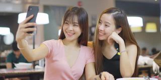 快乐两个年轻的亚洲女孩在餐厅用餐后一起用手机自拍。女人用智能手机看摄像头，然后捕捉到微笑。