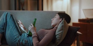 亚洲女性坐在家里的数字平板电脑沙发上喝啤酒