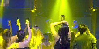 党音乐会。人们用智能手机为社交媒体拍摄照片和视频直播。人群男女在地板上跳舞，音乐家在舞台上表演，灯光效果。