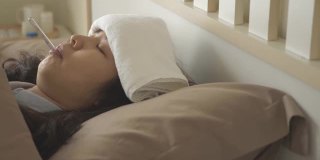 侧视图，摇摄生病的亚洲妇女躺在床上，用毛巾在额头上减轻发烧和降低体温，与口腔温度计在她的嘴里