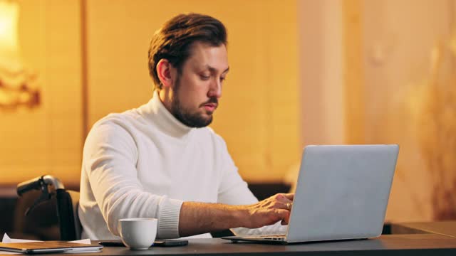 专注的商人企业家在轮椅上打字在笔记本电脑上做研究。年轻男性专业人士坐在家里的办公桌上使用电脑。忙碌的工人自由职业者在现代科技笔记本设备上工作