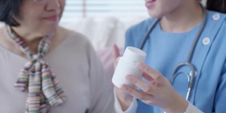 在家访护理服务中，年轻的护理人员穿着手术服向年长的亚洲妇女展示药瓶。亚洲老年人与辅助生活药物监测坐在家里交谈。