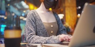 穿着休闲服装的美丽亚洲女人，戴着防护口罩，以防止病毒Covid-19健康危机的传播，在咖啡店使用笔记本电脑工作