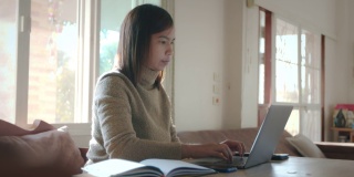 年轻的亚洲女子在电脑上打字，并收到来自智能手机的信息与微笑。在家工作的女性员工。