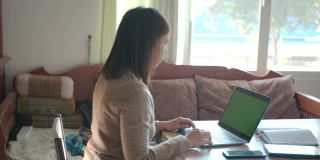 一名年轻的亚洲女子在家里的绿屏笔记本电脑上向参加网络会议的同事问好。在家工作的女性员工。