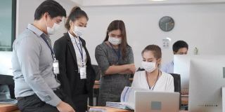 一群年轻的亚洲商人在办公室里用电脑工作，以新的正常生活方式。男男女女因感染新冠肺炎戴上防护口罩，在团队合作中进行头脑风暴和分享想法。