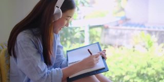 年轻的亚洲女子戴着耳机在笔记本上写东西，然后坐在玻璃窗前