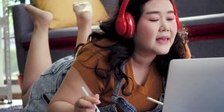 身材高大的女人使用笔记本电脑在家里工作和亚洲女孩喜欢在家里的沙发上吃食物