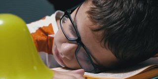 戴眼镜的好奇男孩在桌上看书时睡着了。疲劳、睡眠