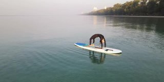 无人机航拍，早晨在SUP Board (Stand - Up Paddle Board)户外锻炼女性瑜伽伸展身体。早晨在阳光柔和的海滩上练习瑜伽。