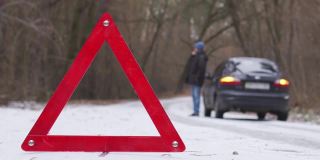 在冬天的森林里，一名男子站在路边一辆带着应急灯的破车旁。红色警告三角形特写。