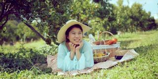 年轻的亚洲迷人的女性穿着休闲服装坐在放松野餐在橙新鲜的花园农场，女孩在野餐与三明治，水果和饮料。农场女孩吃午饭。