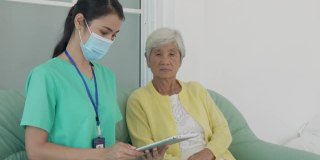 亚洲护士在家客厅用平板电脑对老年患者进行咨询和跟踪护理，在进行家庭保健时在家与医生进行健康咨询和医疗报告。