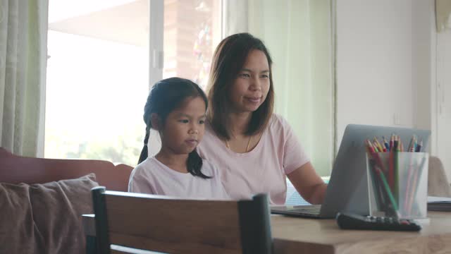 亚洲母女在家里的电脑上通过视频电话问候朋友。由于Covid - 19大流行，在隔离期间儿童在家上学，父母在家工作。