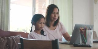 亚洲母女在家里的电脑上通过视频电话问候朋友。由于Covid - 19大流行，在隔离期间儿童在家上学，父母在家工作。