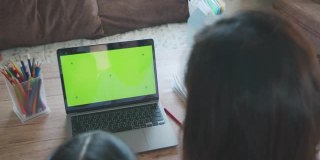 亚洲母女在家里用绿色屏幕的笔记本电脑在线视频通话问候朋友。由于Covid - 19大流行，在隔离期间儿童在家上学，父母在家工作。