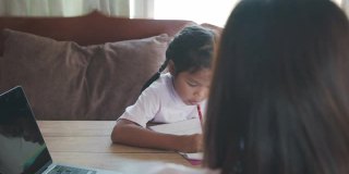 亚洲女童学生做她的家庭作业，而母亲在家工作。由于Covid - 19大流行，在隔离期间，孩子在家上学，父母用电脑在家工作。