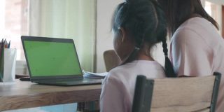 亚洲母女在家里用绿色屏幕的笔记本电脑在线视频通话问候朋友。由于Covid - 19大流行，在隔离期间儿童在家上学，父母在家工作。