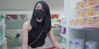 亚洲年轻女性黑色防护面罩推着购物车沿着杂货店通道，妇女在锁定期间得到供应