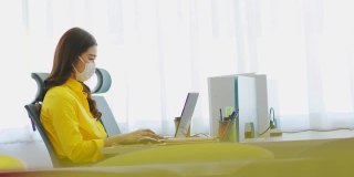 在现代办公室或联合办公空间创业的亚洲女性穿着黄色衬衫，戴着外科口罩，与电脑笔记本思考以获得想法和需求的新常态