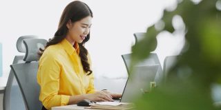 幸福迷人的亚洲女人在黄色衬衫喝咖啡与电脑笔记本思考得到想法和需求在现代办公室或联合办公空间创业
