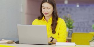 年轻快乐的亚洲商务女性穿着黄色衬衫，与她的业务团队在现代办公室或联合办公空间进行在线视频电话会议