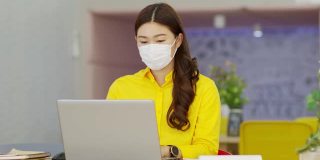 穿着黄色衬衫戴着外科口罩的亚洲女性的新常态通过VDO会议展示销售报告并在图中解释在现代办公室或联合办公空间创业