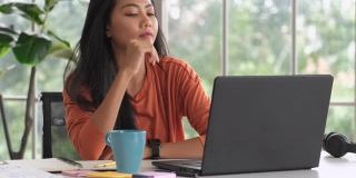 亚洲女性在家工作与微笑和喝咖啡，而工作与笔记本电脑。新常态的生活方式