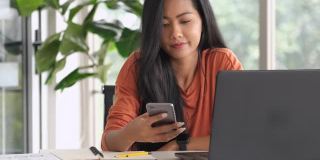 一名亚洲女性在家用笔记本电脑工作时用手机发短信。新常态的办公室生活方式