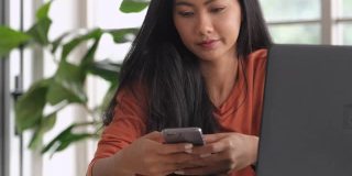 一名亚洲女性在家用笔记本电脑工作时用手机发短信。新常态的办公室生活方式